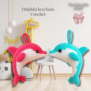 Puede incluir: Se muestran dos llaveros de delfines de ganchillo, uno rosa y blanco, el otro turquesa y blanco. Los llaveros tienen ojos negros y un lazo para colgar. El texto dice "Dolphin keychain Crochet" y "Crochet pattern PDF".