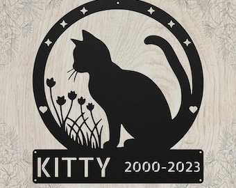 May include: A black metal wall sign featuring a circular frame with stars and hearts. Inside the circle is a silhouette of a cat sitting among flowers. Below the circle is a rectangular plaque with the text KITTY and the years 2000-2023.