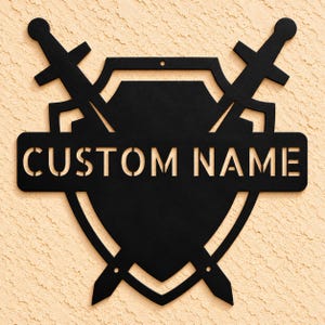 May include: A black metal wall sign featuring a shield emblem centered behind two crossed swords. A horizontal rectangular bar overlays the shield and swords, with the words CUSTOM NAME cut out in a bold, sans-serif capital letter font.