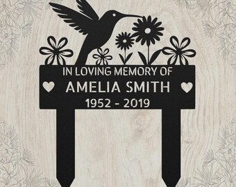 May include: A black metal memorial garden stake featuring a hummingbird silhouette, flowers, and two hearts. The text reads: IN LOVING MEMORY OF AMELIA SMITH 1952 - 2019. The design includes two pointed stakes at the bottom for insertion into the ground.