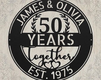 May include: A circular black metal sign featuring the text JAMES & OLIVIA at the top, 50 YEARS in the center, and Together EST. 1975 at the bottom. The design includes two small heart outlines and laurel branch accents within the circular frame.