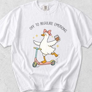 Puede incluir: Una camiseta blanca de manga corta con el gráfico de una oca blanca con un lazo rosa, montada en un patinete rosa y azul mientras sostiene un vaso de café. El texto sobre la oca dice "OFF TO REGULATE EMOTIONS" junto a pequeños iconos de estrellas.