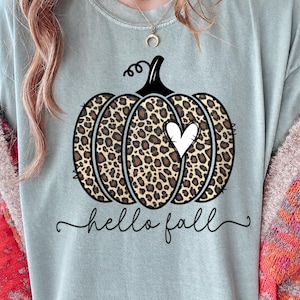 May include: A sage green t-shirt featuring a central graphic of a pumpkin with a brown and black leopard print pattern. The pumpkin has a black stem and a small white heart shape on its side. Below the pumpkin, the text hello fall is written.