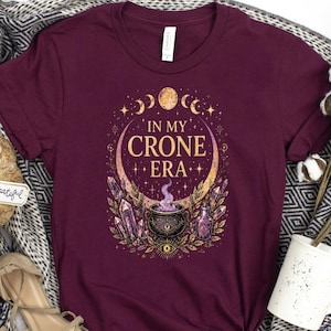 May include: A maroon short-sleeved t-shirt featuring a graphic print with the text "IN MY CRONE ERA" surrounded by celestial moon phases, a steaming cauldron, crystals, and botanical elements in shades of gold, purple, and white.
