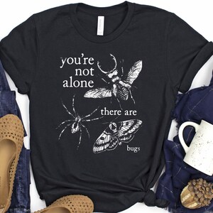 Peut inclure: Un t-shirt noir à manches courtes avec une impression graphique blanche représentant un scarabée, une araignée et un papillon de nuit avec le texte "you're not alone there are bugs". Il est accompagné d'un jean bleu foncé, de ballerines et d'une écharpe.