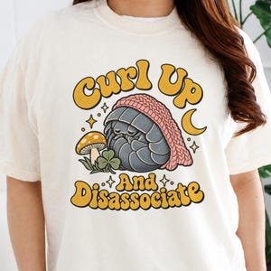 May include: A cream-coloured t-shirt featuring a graphic of a grey pill bug wearing a pink knit hat, accompanied by a mushroom and a clover. The text reads "Curl Up And Disassociate" in a bold, yellow, rounded font with black outlines.