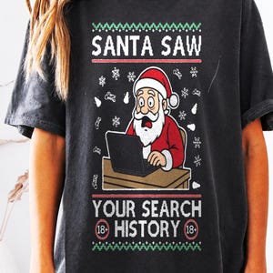 Puede incluir: Una camiseta gris carbón de manga corta con un gráfico de Santa Claus mirando sorprendido un portátil. El texto dice SANTA SAW YOUR SEARCH HISTORY con símbolos de 18+. El diseño incluye patrones de copos de nieve y bordes rojos y verdes.