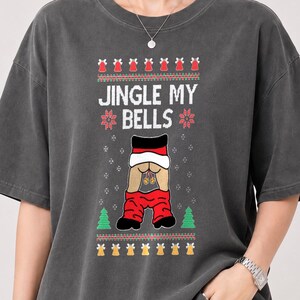 May include: A charcoal grey short-sleeve t-shirt featuring a graphic print with the text JINGLE MY BELLS. The design includes red and green holiday patterns, a pair of trousers pulled down, and two small gold bells hanging from the centre area.