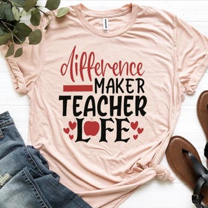 Puede incluir: Una camiseta de manga corta de color rosa claro con el texto en rojo y negro "Difference MAKER TEACHER LIFE", un gráfico de una manzana roja que sustituye a la letra A en "LIFE" y varios corazones rojos pequeños alrededor.