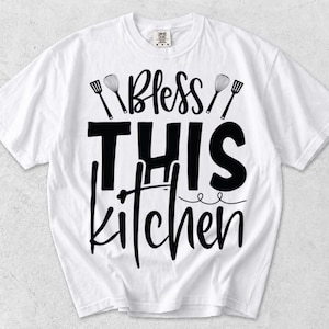 以下が含まれることがあります： Bless THIS kitchenという黒い文字がプリントされた白い半袖Tシャツ。デザインには、衣服の前面の文字の上と周囲に配置された2つの泡立て器と2つのスパチュラのグラフィックイラストが含まれています。