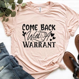 May include: A light pink short-sleeved t-shirt featuring black text that reads "COME BACK With A WARRANT" in a mix of bold and script fonts, accented with small black heart icons and decorative flourishes centred on the chest.