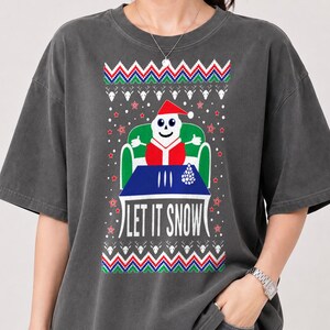 May include: A charcoal grey t-shirt featuring a graphic print of a white skull character wearing a red Santa hat and suit, sitting in a green chair behind a blue table. The text "LET IT SNOW" is printed below, surrounded by festive patterns.