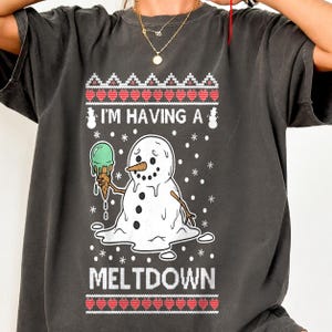 Puede incluir: Una camiseta gris carbón con un gráfico de un muñeco de nieve derritiéndose que sostiene un helado verde. El texto dice I'M HAVING A MELTDOWN. El diseño incluye copos de nieve blancos y un borde con patrón rojo y blanco en la parte superior e inferior.