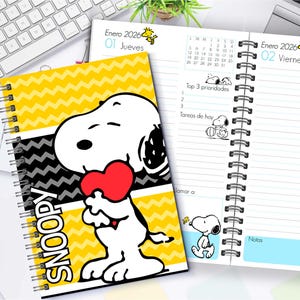 May include: A spiral-bound Snoopy planner with a yellow and black chevron pattern cover. The cover features Snoopy holding a red heart. The open planner shows daily entries with Snoopy illustrations and the text "Enero 2026".