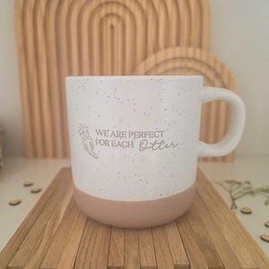 Könnte beinhalten: Weiß gesprenkelter Keramikbecher mit hellrosa Boden und gebogenem Henkel. Der Becher trägt den Text "WE ARE PERFECT FOR EACH Otter" mit einer Otter-Illustration. Der Becher steht auf einer Holzoberfläche.