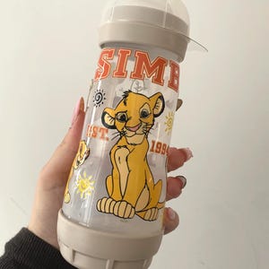 Peut inclure: Un biberon transparent avec un motif Simba du Roi Lion. Le biberon a une base et un dessus beiges, avec le nom "SIMBA" en lettres orange. Le motif comprend un lionceau jaune, des soleils et l'année "1994".