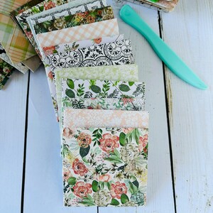 May include: A stack of patterned paper squares with floral and geometric designs in shades of green, white, and pink. The papers are arranged in a fan shape on a light blue surface.