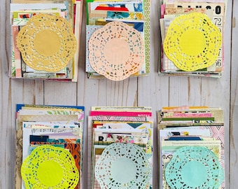 May include: Six stacks of paper ephemera with colorful patterned papers, vintage ephemera, and decorative doilies in yellow, peach, and green. The stacks are arranged on a light wood surface.