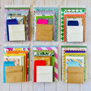 May include: Six stacks of assorted stationery items, each featuring a variety of colorful envelopes and patterned note cards. The collection includes solid-colored, floral, checkered, and striped designs in various sizes, layered to display multiple textures.