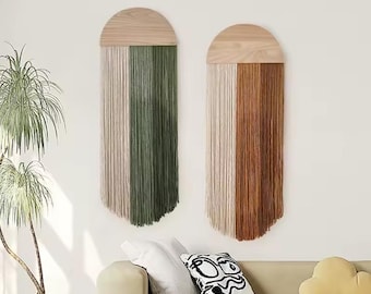 May include: Two wall hangings with a wooden half-circle top and vertical fringe. One features green and beige fringe, the other brown and beige. The decor is mounted on a white wall, with a sofa and pillows in the foreground.