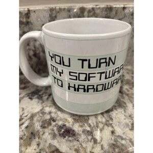 May include: White ceramic mug with a curved handle. The mug features the text "YOU TURN MY SOFTWARE TO HARDWARE" in black block letters, with light green stripes. The mug is on a speckled surface.