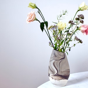 Peut inclure: Un vase en céramique marbré gris et blanc avec une forme asymétrique unique, contenant un bouquet de fleurs roses et blanches avec des tiges vertes.