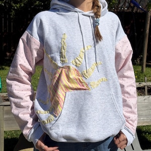 May include: A gray hoodie with a sun design. The sun is made of yellow fabric and pink and yellow striped fabric. The sleeves are pink with black polka dots. The hoodie has a front pocket and a hood with drawstrings.
