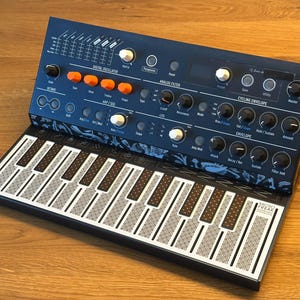 Könnte beinhalten: Ein dunkelblauer Arturia MicroFreak Synthesizer mit einer flachen, berührungsempfindlichen Tastatur mit weiß-schwarzer Musterung. Das Bedienfeld enthält orangefarbene Knöpfe, schwarze Drehregler und weiße Tasten mit Beschriftungen für Digitaloszillator, Analogfilter, Cycling Envelope und Arp/Seq-Funktionen.