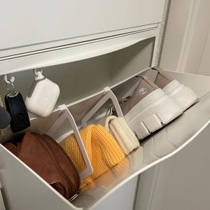 Könnte beinhalten: Ein weißes Aufbewahrungsmodul mit mehreren Fächern und einer herunterziehbaren Tür. Im Inneren befinden sich Schlüssel, ein Autoschlüssel und ein weißes kabelloses Ladecase, die an Haken hängen. Außerdem eine braune Handtasche, gelbe und cremefarbene Mützen und weiße Plateauschuhe.