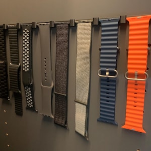 May include: A collection of various smartwatch bands in different colors and styles, including black metal, black perforated, gray, dark gray, light gray and orange silicone bands. The bands are displayed on a black rack.