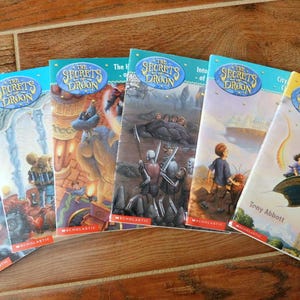 May include: A set of seven paperback books with blue and white covers featuring illustrations of dragons and fantasy scenes. The books are titled "The Secrets of the Dragon" and each book has a different subtitle.
