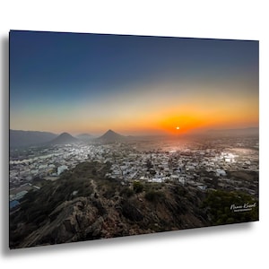 May include: A landscape photograph of a city at sunset. The sky transitions from blue to orange, with the sun setting over the horizon. The city is nestled between mountains, with a rocky foreground.