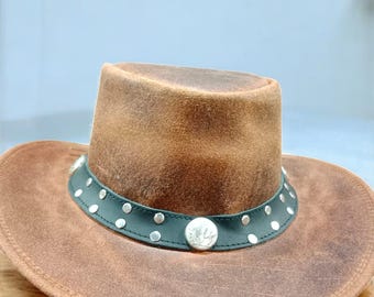 Könnte beinhalten: Ein brauner Cowboyhut mit einem Leder-Look. Der Hut hat ein dunkelgrünes Band mit silberfarbenen Nieten und einem großen silbernen Emblem. Der Hut liegt auf einer Holzoberfläche.