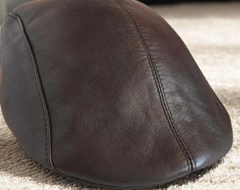 May include: A brown leather flat cap with a rounded crown and a small, stiff brim. The cap has a smooth, glossy finish and is stitched along the seams. The hat is resting on a light-colored, textured surface.