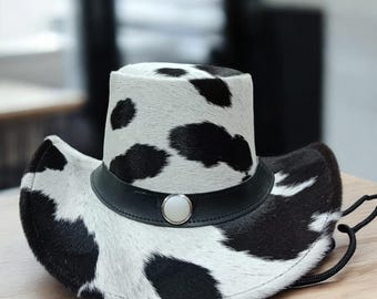 May include: Cowboy hat with a black and white cowhide pattern. The hat has a black leather band with a silver-coloured button and a black chin strap. The hat is on a wooden surface.