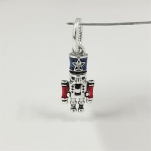 May include: A silver charm shaped like a toy soldier nutcracker. The nutcracker has a blue and silver hat with a star and a clear gem. The arms are red, and the body is silver. The charm is suspended from a silver loop.