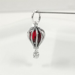 Può includere: Un charm d'argento a forma di mongolfiera con un centro rosso. Il charm presenta un design a gabbia dettagliato e un piccolo anello nella parte superiore per il fissaggio a un braccialetto o una collana. La parola "Pandora" è incisa sul fondo del charm.
