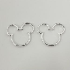 Puede incluir: Dos pendientes de aro plateados con forma de la cabeza de Mickey Mouse. Los pendientes tienen un acabado liso y pulido y la palabra "PANDORA" está grabada en la parte superior de cada pendiente. Se muestran sobre un fondo blanco.