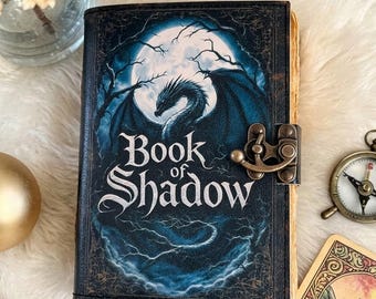 May include: A rectangular journal titled Book of Shadow with a dark blue cover featuring a dragon silhouette against a full moon and bare tree branches. The book has a metallic bronze-colored latch closure and visible aged paper pages on the side.