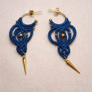 Peut inclure: Une paire de boucles d'oreilles macramé bleues faites à la main avec des accents dorés. Les boucles d'oreilles présentent un motif tissé avec des perles dorées et un pendentif en forme de pointe dorée. Les boucles d'oreilles sont sur un fond clair.