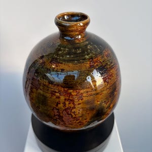 Puede incluir: Un jarrón de cerámica hecho a mano con un cuerpo redondeado y un cuello estrecho. El jarrón presenta una mezcla de tonos marrón, óxido y negro, con un acabado brillante. La textura parece tener un veteado similar a la madera. El jarrón se exhibe sobre un soporte negro.