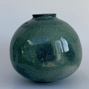 Può includere: Un vaso in ceramica color teal con corpo rotondo e collo stretto. Il vaso presenta una finitura craquelé, che gli conferisce un aspetto strutturato. La superficie riflette la luce, evidenziando le variazioni di colore. Circa 20 cm di altezza.