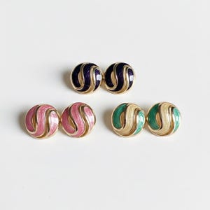 May include: Three pairs of vintage stud earrings in gold-tone metal. One pair is navy blue, another is pink, and the third is green and cream. Each earring features a swirling design, perfect for adding a pop of color to any outfit.