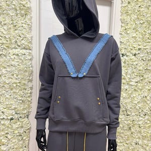 May include: A dark gray hoodie and matching sweatpants set displayed on a mannequin. The hoodie features a blue denim V-shaped accent across the shoulders, gold-colored rivets, and a front pocket. The sweatpants have gold drawstrings.