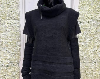 May include: A black, textured sweater with a high, draped collar and a silver chain detail. The sweater features short sleeves and attached arm warmers. The mannequin is wearing black pants. The background is a white wall with white floral decorations.
