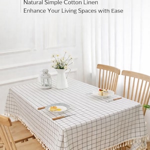 May include: White cotton linen tablecloth with a black grid pattern and fringe trim. The text on the image reads: "Elegant Cotton Linen Tablecloth, Natural Simple Cotton Linen, Enhance Your Living Spaces with Ease."