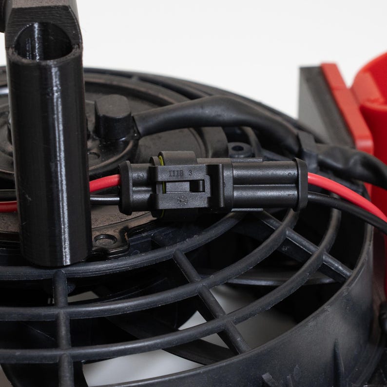 May include: Close-up of a black electrical connector with red and black wires, connected to a black fan. The fan has a black plastic housing and blades. A black tube is visible on the left side of the image. The background is white.