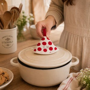 Puede incluir: Una olla de cerámica color crema con tapa, coronada por un agarrador de tela triangular con lunares rojos y las iniciales K.M. bordadas. El agarrador tiene una pequeña presilla de tela roja en la parte superior para colgarlo fácilmente.