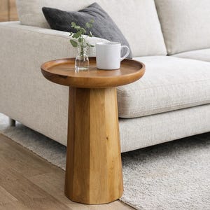 May include: A small, round wooden side table with a pedestal base. The tabletop holds a white mug, a small glass vase with greenery, and a grey cushion rests on a light-coloured sofa. The table is made of light-coloured wood.