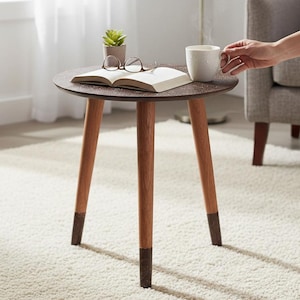 Może przedstawiać: Mały, okrągły stolik pomocniczy z ciemnobrązowym, nakrapianym blatem i trzema drewnianymi nogami. Nogi są zaakcentowane ciemniejszym brązem u podstawy. Na stole książka, okulary, mała roślina i kubek.